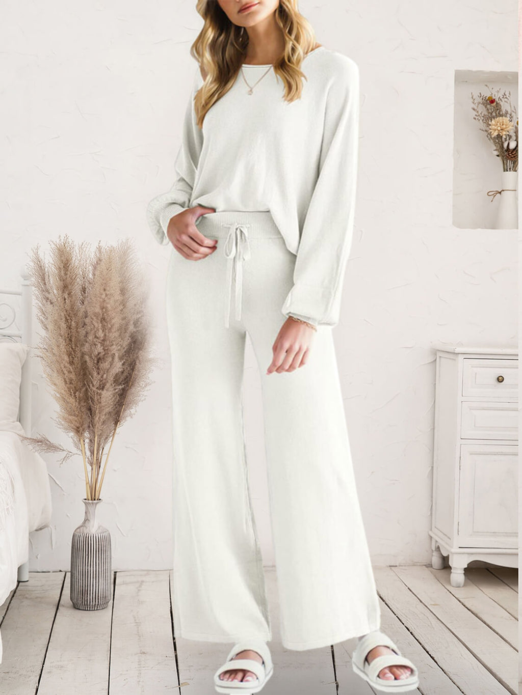 A woman models a matching two-piece lounge set consisting of a long-sleeve crewneck top and high-waisted wide-leg pants, both in a soft off-white/cream color. The set is styled in a bright, neutral bedroom setting with a wooden floor, a white dresser, and a vase of pampas grass.