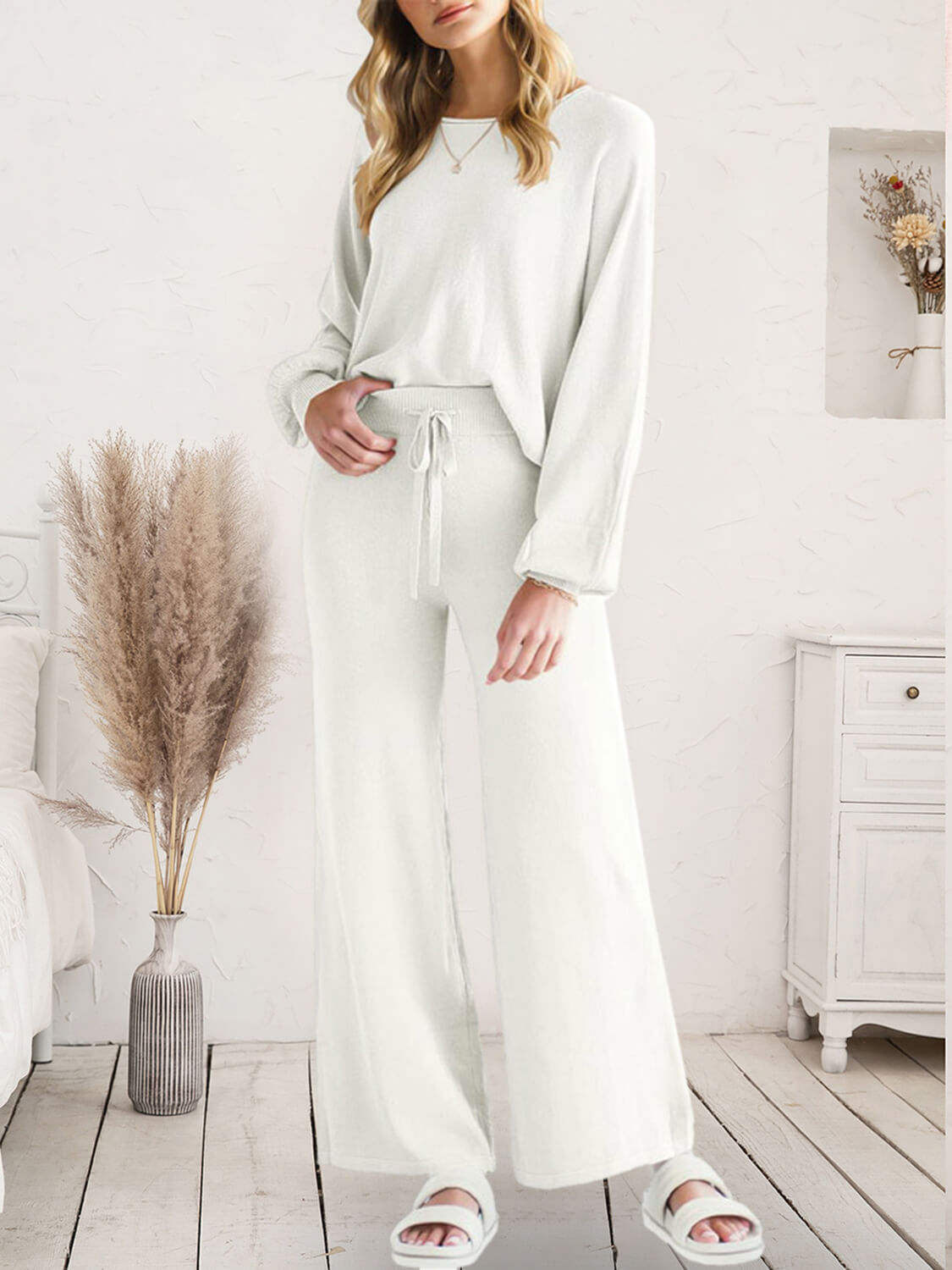 A woman models a matching two-piece lounge set consisting of a long-sleeve crewneck top and high-waisted wide-leg pants, both in a soft off-white/cream color. The set is styled in a bright, neutral bedroom setting with a wooden floor, a white dresser, and a vase of pampas grass.