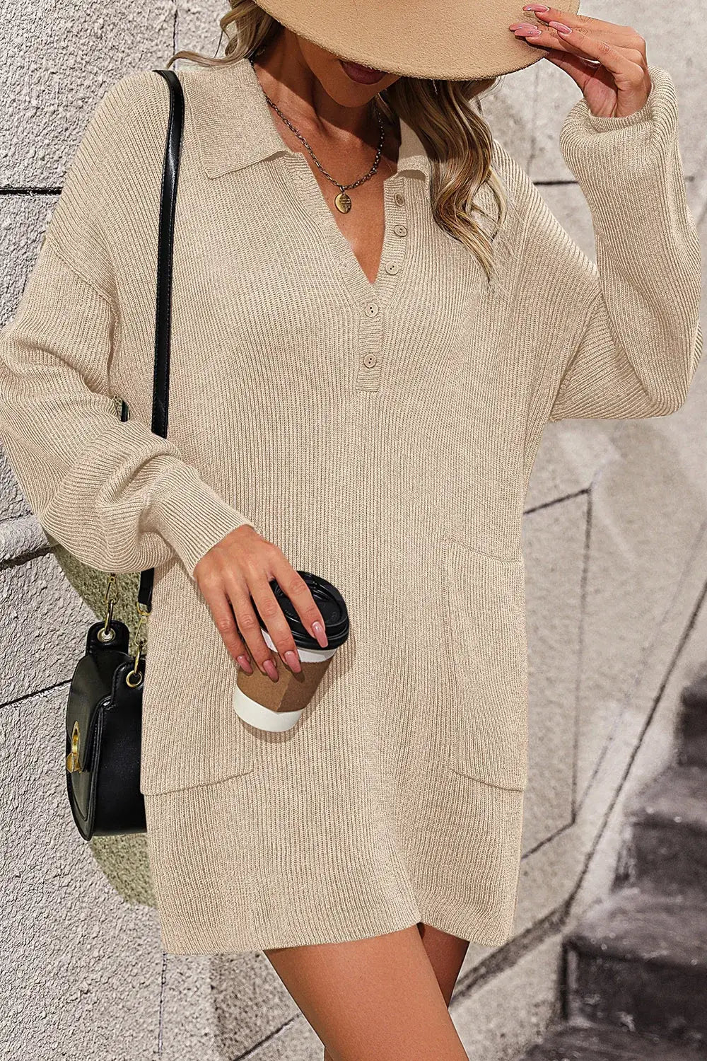 Woman wearing a beige sweater dress holding a coffee cup against a textured wall.
