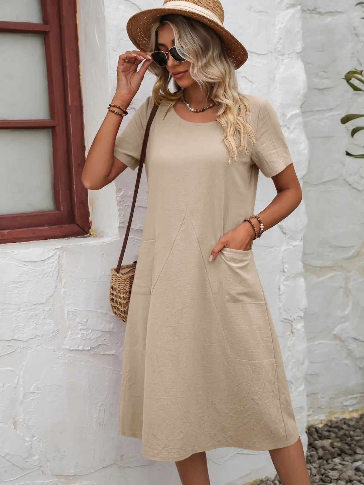 Woman wearing a beige dress and straw hat against a white stone wall.