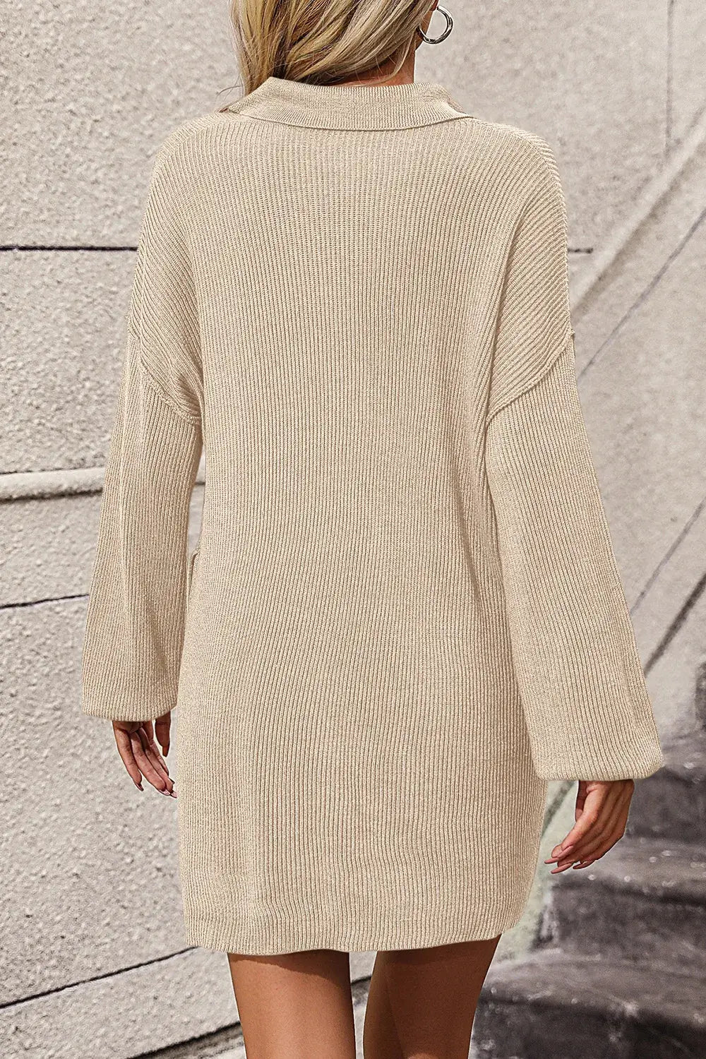 A woman wearing a beige ribbed knit sweater dress, shown from the back against a textured concrete wall.